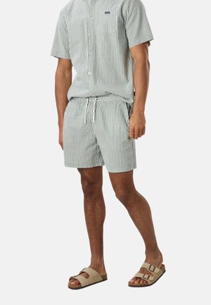 Mężczyzna ubrany w szaro-białą, prążkowaną koszulę z krótkim rękawem na guziki oraz pasujące szorty ze sznurkiem, beżowe sandały z podwójnymi paskami.