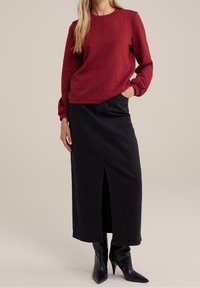 Roter strukturierter Pullover mit langen Ärmeln, kombiniert mit einem schwarzen Jeans-Maxirock mit einem vorne eingeschnittenen Schlitz, ergänzt durch schwarze spitze Stiefel.