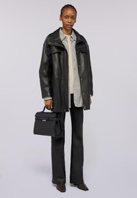Giubbotto di pelle nera con vestibilità oversize, chiusura a bottoni, due tasche frontali, indossato sopra una camicia a righe e un maglione beige, abbinato a pantaloni a zampa d'elefante e borsa.