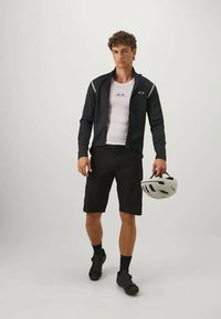 Νεαρός άνδρας με μαύρη ζακέτα ποδηλασίας, λευκό πουκάμισο, μαύρες βερμούδες και παπούτσια, κρατάει ένα λευκό κράνος ποδηλασίας μπροστά από απλό φόντο.