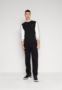 Felpa nera con maniche grigie e bianche, dotata di scollo a giro e piccolo logo verde. Il modello indossa pantaloni neri e sneaker nere.