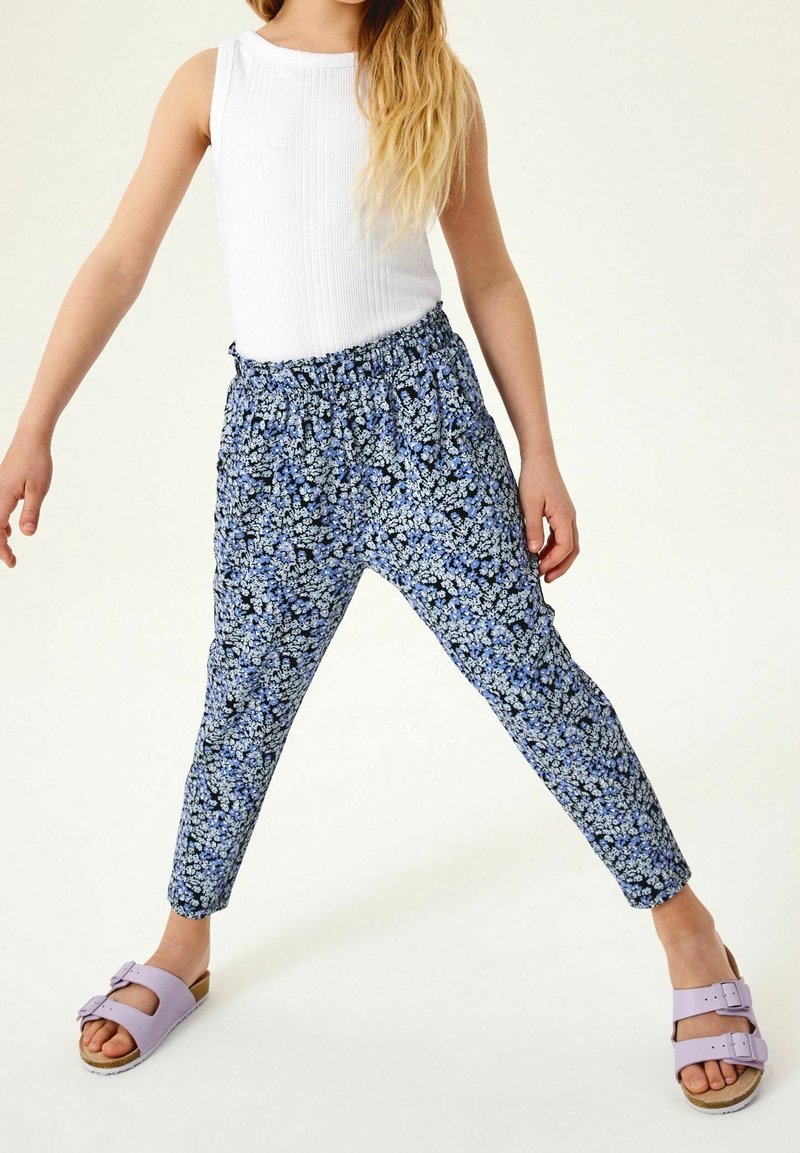 Enfant portant un haut blanc sans manches, un pantalon bleu à motif floral et des sandales lavande avec boucle, debout, jambes écartées.