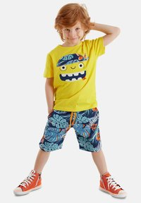 Camiseta de algodón amarilla con diseño de cara de monstruo, combinada con pantalones cortos azules con un patrón de hojas tropicales y cordón naranja, y zapatillas deportivas naranjas.