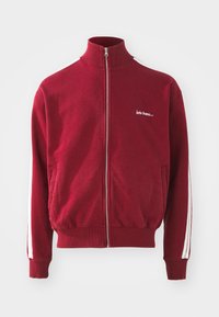 Punane lukuga spordijakk kõrge kaelusega, valgete külgtriipudega ja vasakul rinnal brodüüritud tekstiga. Tekst on valge. Valmistatud pehmest kangast.