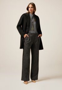 Schwarzer Mantel über einem grauen Pullover, kombiniert mit grauen, strukturierten, weit geschnittenen Hosen. Beigefarbene Absatzschuhe und eine lange, hängende Halskette runden das Outfit ab.