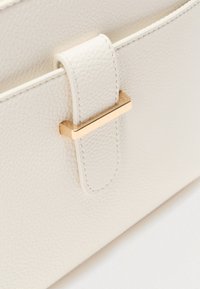 Λευκή τσάντα από δέρμα με υφή, με επίχρυσο ορθογώνιο μεταλλικό διακοσμητικό και χοντρό λουράκι. Διαθέτει λεία υφή και καθαρές γραμμές.