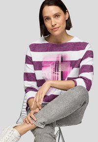 Lila- und weiß gestreifter Pullover mit Grafikun Gestaltung. Der Stoff wirkt strukturiert. Das Model sitzt mit grauen Hosen und weißen Schuhen.