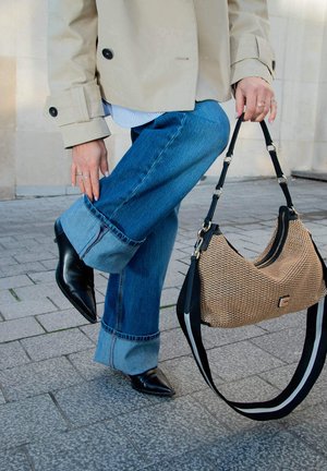 Persoon in opgerolde blauwe spijkerbroek, beige jas en zwarte laarzen die een beige geweven handtas met zwarte handvatten en een gestreepte schouderband buiten vasthoudt.