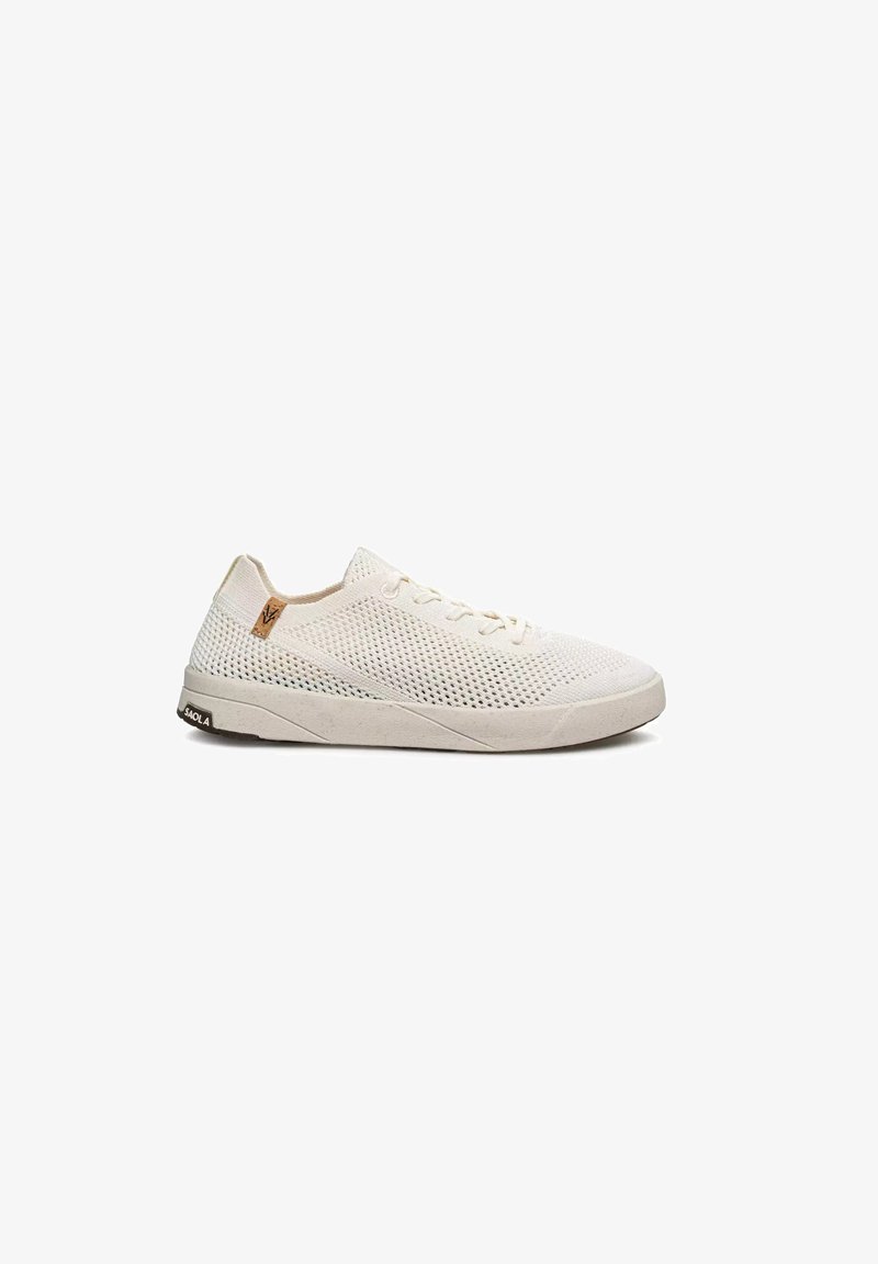 Zapatilla deportiva de perfil bajo color beige claro con parte superior de malla, cierre de cordones, suela plana de goma y pequeña etiqueta con logo marrón cerca del talón.