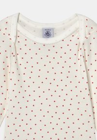 Langärmliger Bodysuit aus weichem cremefarbenem Baumwollstoff, mit einem zarten roten Herzdruck, gewelltem Ausschnitt und einem Etikett zur Größenangabe.