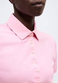 Πρόσωπο με σκουρόχρωμο δέρμα που φοράει ένα ανοιχτό ροζ πουκάμισο με γιακά και έξι κουμπιά σε μια απλή ανοιχτή φόντο.