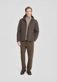 Giacca imbottita marrone con cappuccio, indossata sopra un dolcevita beige. Abbinata a pantaloni marroni e scarpe scure. Design semplice e pulito.