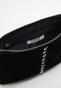Clutch de ante negro con tachuelas en forma de pirámide plateadas, cremallera abierta mostrando el interior de tela negra y etiqueta de la marca "PIECES" dentro.