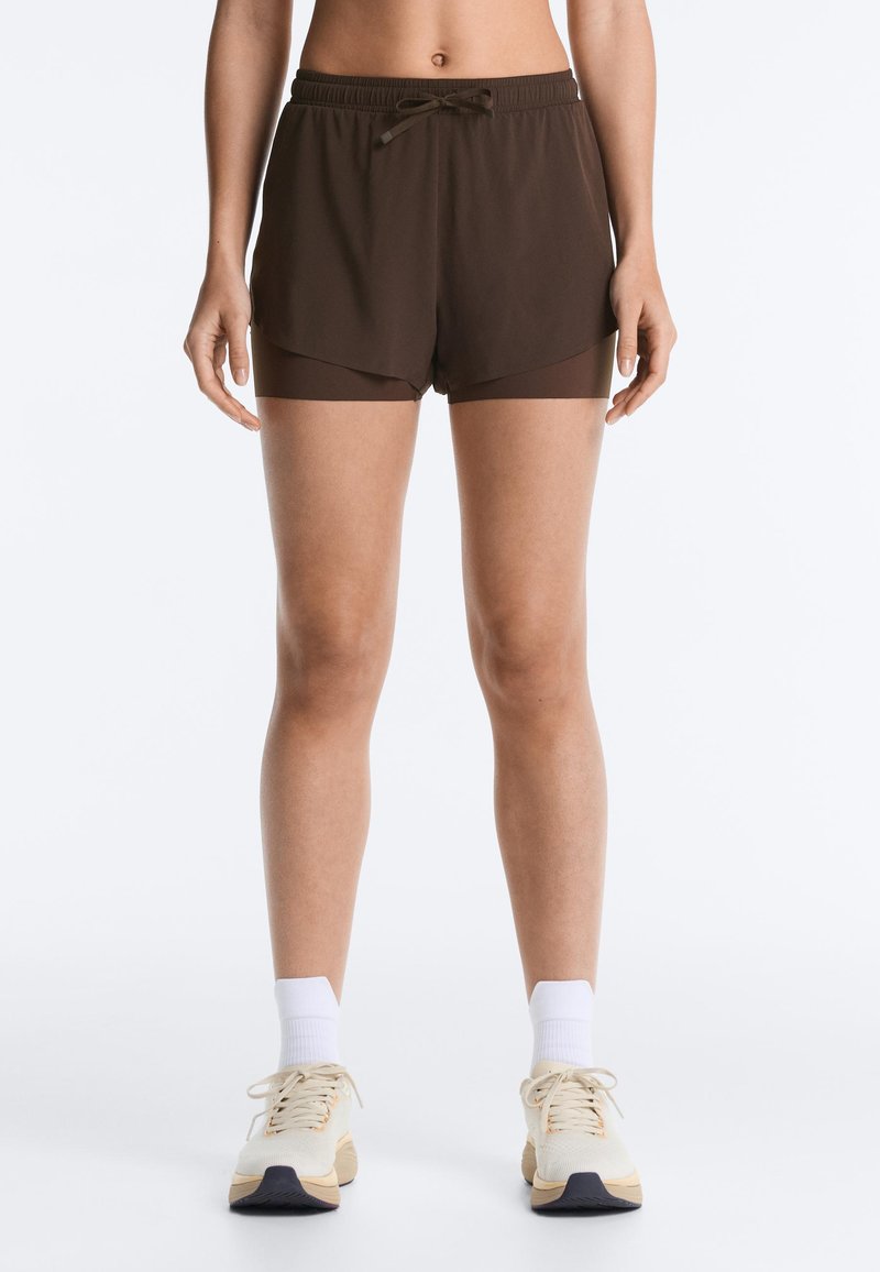 Woman wearing dark brown layered athletic shorts with drawstring, white crew socks, and beige running shoes, standing against white background.