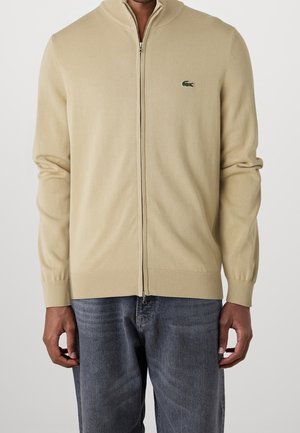 Homme portant un pull beige zippé avec un petit logo crocodile vert et un jean bleu délavé sur un fond uni.