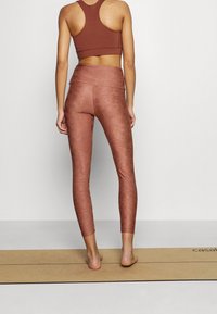 Brązowy stanik sportowy z plecami w stylu racerback, zestawiony z wysokim stanem, strukturalnymi legginsami w kolorze rdzy, z subtelnym wzorem.