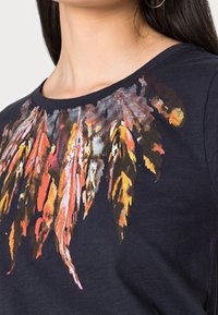 Tummansininen T-paita, jossa on kirkas, monivärinen höyhenkujatus design, joka sisältää oranssin, keltaisen ja vihreän sävyjä kaulan ympärillä. Puuvillamateriaalia.