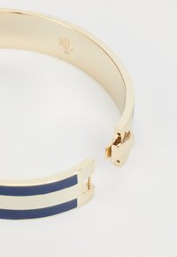 Bracciale in metallo color oro con strisce alternate blu navy e bianche, finitura liscia e chiusura a scatto sicura. Incisione del logo all'interno.