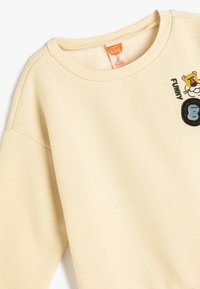 Licht beige sweatshirt met een ronde hals. Heeft een leeuw graphic met de tekst "GRAPPIG" en een ronde blauwe aap patch. Zacht, textielachtig materiaal.