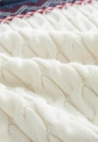 Tissu épais tricoté de couleur crème avec un motif en torsade tressée et un aperçu textile rouge et bleu en arrière-plan.