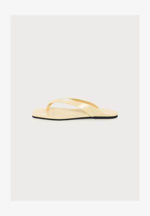 Beige platte teenslipper met gekruiste bandjes en een dunne teenslipperdetail op een zwarte zool, zijaanzicht op een witte achtergrond.