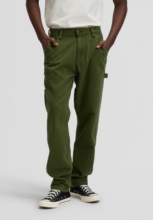 Személy olívazöld cargó nadrágot, fehér inget (betűrve), zsebébe dugott kezekkel és fekete, fehér fűzős alacsony szárú sportcipővel.