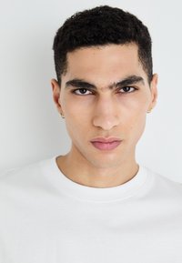 Camiseta blanca de cuello redondo, usada por un modelo masculino con cabello corto, oscuro y rizado. Detalles notables incluyen pendientes de aro dorados y rasgos faciales definidos.