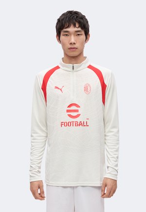 Jeune homme portant un haut d'entraînement de football blanc avec des accents rouges, logo Puma, écusson AC Milan et branding eFootball, debout devant un fond uni.