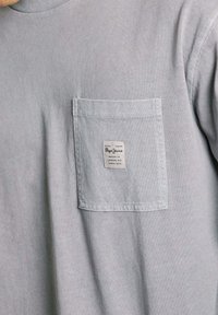 Grau kurzärmliges T-Shirt aus Baumwolle mit einer linken Brusttasche und einem Markenlabel, weichem Material und klassischem Rundhalsausschnitt.