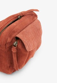 Roestkleurige crossbodytas van getextureerd leer met voorvleugeltasje en hoofdvak met ritssluiting, afgebeeld op een witte achtergrond.