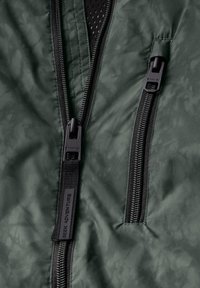 Green jacket featuring two black zippers and textured fabric. Includes a label stating "REEK ADVENTURE" near the zipper area.
