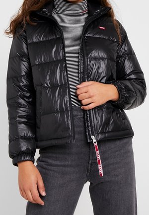 Personne portant une doudoune noire brillante avec le logo Levi's, un col roulé rayé et un jean gris foncé, main saisissant la fermeture éclair de la veste.