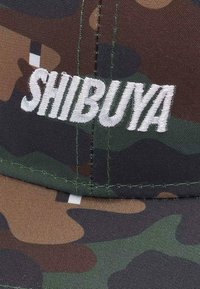 Camo-Muster-Kappe aus Stoff in Grüntönen, Brauntönen und Schwarz; mit weißem, gesticktem "SHIBUYA"-Text auf der Vorderseite.