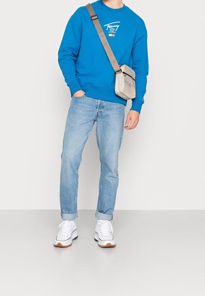 Blauwe sweatshirt met wit logo, lichtblauwe jeans en witte sneakers met een gumrubberen zool. Inclusief een kleine beige schoudertas.