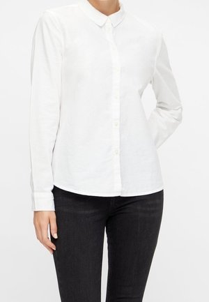 Chemise blanche à manches longues, boutonnée avec col, portée avec un jean noir ajusté sur une personne debout devant un fond uni.