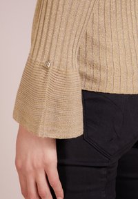 Pull en or à côtes avec des manches évasées, présentant un bouton décoratif en argent au niveau des poignets. Porté avec un jean noir affichant des coutures discrètes.