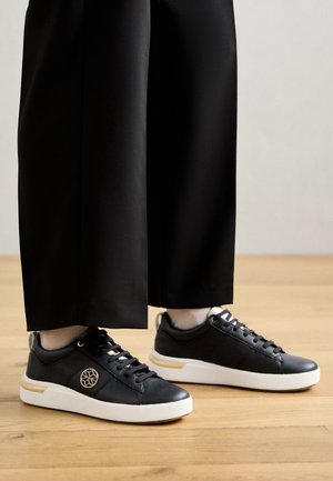 Baskets en cuir noir avec une finition texturée, semelle en caoutchouc blanche et détail en accent doré. Les lacets sont noirs, avec un logo rond sur le côté.