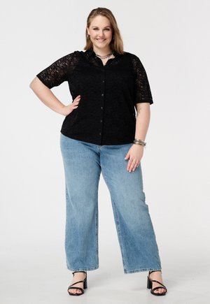 Lachende vrouw staat met hand op heup, draagt een zwart kanten kort mouw bloesje, lichtblauwe wijde jeans en zwarte sandalen met hak.