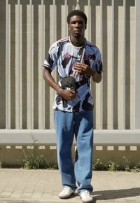 Pánske tričko s krátkym rukávom s modro-čiernym abstraktným vzorom, skombinované so svetlomodrými voľnými džínsami a bielymi teniskami. Malá čierna kabelka cez rameno.