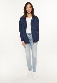Felpa con zip blu navy e cordini, shirt bianca a girocollo, jeans fitted azzurri chiari e sneakers bianche, esposti su un modello in piedi.