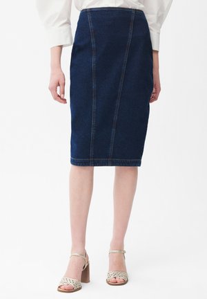 Kobieta ubrana w ciemnoniebieską ołówkową spódnicę dżinsową i białe sandały na blokowym obcasie, stojąca na tle jednolitego białego tła.