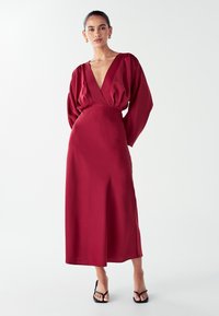 Moteris vilkinti tamsiai raudoną, ilgomis rankovėmis, V formos kaklo midi suknelę su pūstomis rankovėmis ir juodais atvirais bateliais su kulnu, stovinti prieš paprastą baltą foną.