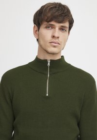 Maglione lavorato a maglia verde scuro con un colletto alto e una zip argentata. Presenta un motivo testurizzato su tutto il tessuto.