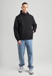 Black hooded jacket with two front pockets, zipper closure, and ribbed cuffs, paired with light blue jeans and white sneakers.
