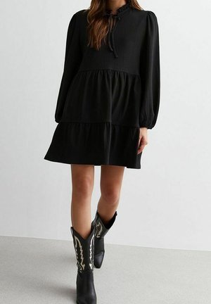 Femme portant une robe mini noire à manches longues et à volants, associée à des bottes de cow-boy noires avec des coutures décoratives blanches.