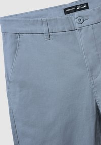 Szare spodnie z bawełnianego chambray z płaskim przodem, szlufkami na pasek i pojedynczym zapięciem na guzik. Kieszenie po bokach i przeszycia na szwach.