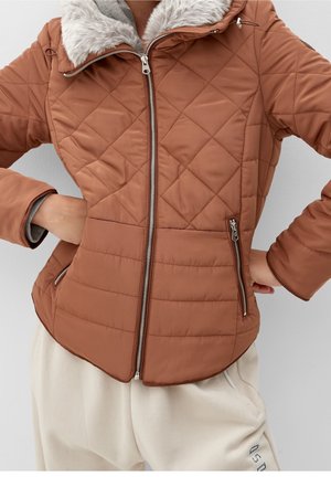 Femme portant une veste matelassée couleur rouille avec un col en fourrure et des poches zippées, associée à un pantalon beige clair.