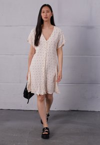 Vestido de bolinhas em bege claro com decote em V, mangas curtas e fluídas, frente de botões e tecido suave e leve; combinado com uma bolsa preta e sandálias robustas.