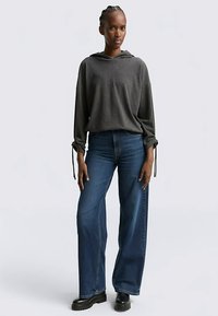 Grijze capuchontrui met koordjes bij de mouwen, gecombineerd met blauwe jeans met brede pijpen en zwarte grofgebouwde laarzen. Gladde stof, losse pasvorm.