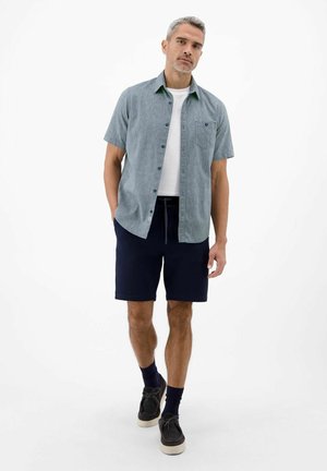 Mężczyzna z siwymi włosami nosi szarą koszulę z krótkim rękawem zapinaną na guziki, białą koszulkę, granatowe szorty, czarne buty i granatowe skarpetki, stoi na białym tle.